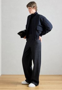 Tumši zila bumbvedēja jaka ar izšūtu rakstu, divām priekšējām kabatām, kopā ar augsta vidukļa tumši zila džinsa biksēm un baltām kedām. Rokā tiek turēts melns makiņš.