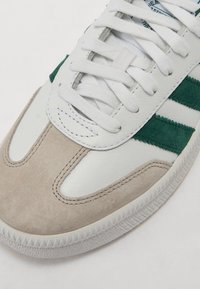 Hvite sneakers med grønne semskede striper og en beige semsket tå. Teksturet hvit skinnkropp med flate hvite lisser og gummisåle.