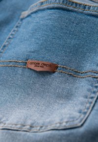 Tissu en denim de teinte bleu clair, avec une étiquette en cuir brun embossée des inscriptions « ombre dept » et « since 1985 » cousue sur la poche.