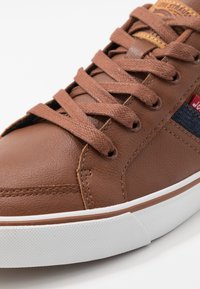 Brązowy skórzany tenisówka z teksturowaną powierzchnią, zaokrąglonym noskiem i kontrastującym panelem z materiału. Posiada brązowe sznurowadła, metalowe przelotki i białą podeszwę.