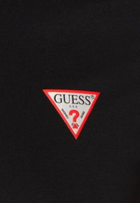 Musta kangas, jossa on punaisella reunustettu valkoinen kolmion muotoinen Guess-logo, jossa on punainen kysymysmerkki ja teksti "Yhdysvallat. Pestyt farkut."