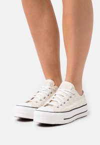 Sneakers w kolorze kremowym wykonane z płótna, z wzorowaną fakturą, białymi gumowymi podeszwami, czarnymi paskami i srebrnymi oczkami na sznurówki.