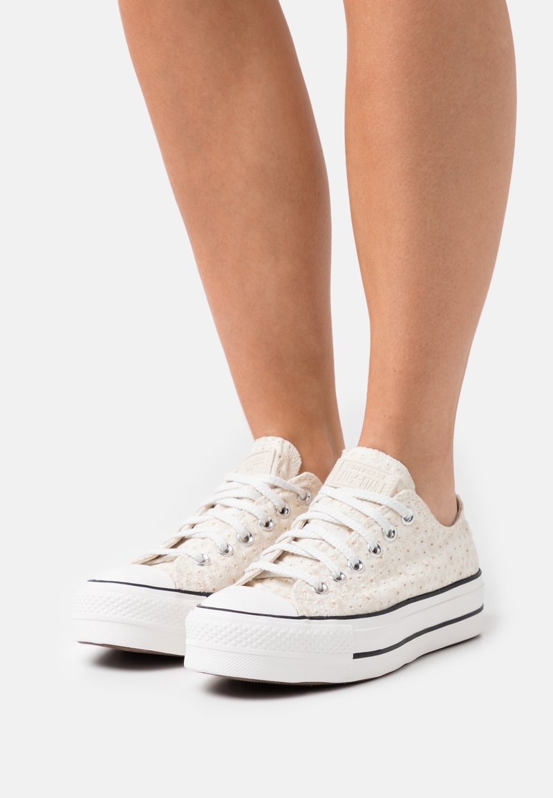 Sneakers w kolorze kremowym wykonane z płótna, z wzorowaną fakturą, białymi gumowymi podeszwami, czarnymi paskami i srebrnymi oczkami na sznurówki.