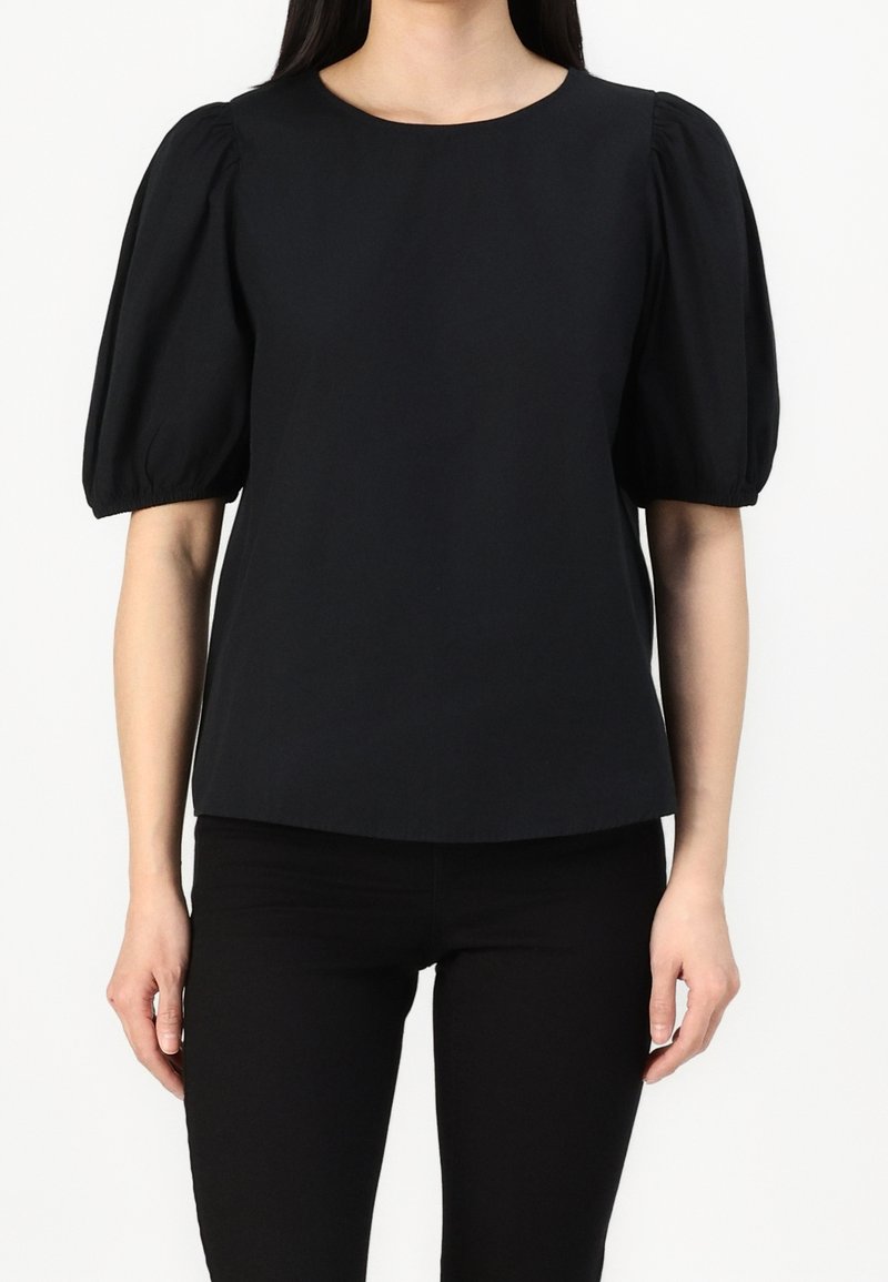 Femme portant un chemisier noir avec un col rond et des manches courtes bouffantes, associé à un pantalon noir, debout devant un fond blanc.