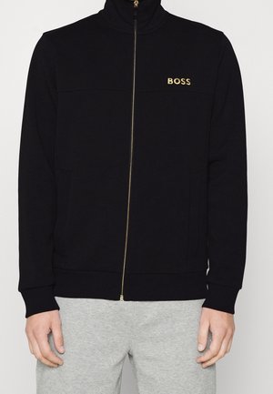 Sudadera negra con cremallera y cuello alto, logo dorado "BOSS" en el pecho, puños y dobladillo acanalados, y dos bolsillos frontales. Textura de tela suave.