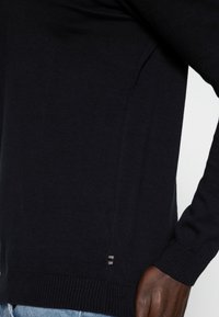 Pull en maille bleu marine avec une coupe ajustée, des poignets côtelés et une subtile fente latérale. Texture douce avec une finition lisse.