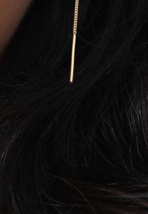 Boucle d'oreille en chaîne dorée avec une fine barre verticale suspendue à côté de longs cheveux noirs ondulés.