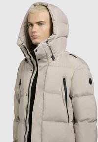 Chaqueta de plumas beige con una gran capucha, cremalleras negras a los lados y una textura suave con relleno acolchado.