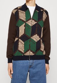 Stickad jacka med dragkedja i brunt med ett geometriskt mönster i grönt, beige och marinblått. Har en ribbad krage och muddar.