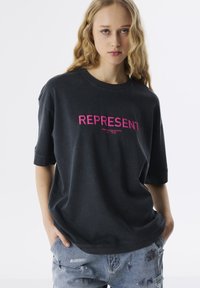 Black oversized t-shirt featuring bold pink "REPRESENT" text on the front. Short sleeves and a relaxed fit complement distressed blue jeans.