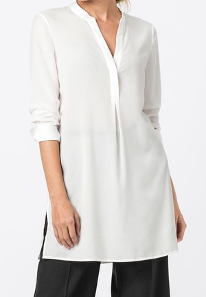 Femme portant une blouse tunique blanche à manches longues avec un col mandarin, une fente en V au col et des manches retroussées, assortie à un pantalon noir.