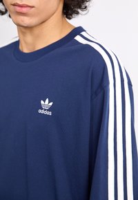 Mørkeblå bomulds t-shirt med hvidt tre-stribe design på ærmerne og lille hvid Adidas logo på brystet. Casual fit, rund hals.