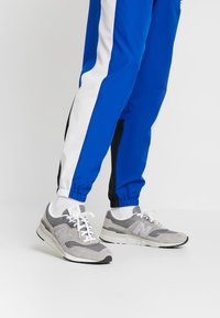 Grå atletiska skor med texturerad mocka och mesh, med vita snören och en framträdande 'N'-logotyp, i kombination med blå joggers.