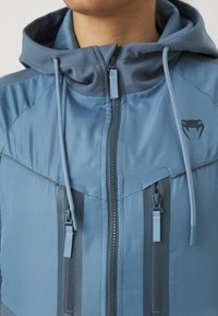 Casaco azul com capuz e acabamento texturizado, apresentando um fecho frontal com zíper, cordões ajustáveis e dois bolsos com zíper; logótipo no lado superior esquerdo.