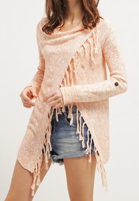 Cardigan en maille pêche clair avec devant drapé, bords effilochés, manches longues et bouton décoratif au poignet. Associé à un short en denim.