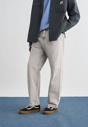 Persona che indossa jeans grigio chiaro a vestibilità rilassata, sneakers nere e bianche con suole marroni, camicia azzurro chiaro e giacca scura con bottoni.