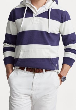 Uomo che indossa una felpa con cappuccio a strisce blu navy e bianche, pantaloni bianchi, cintura intrecciata marrone e orologio da polso con quadrante blu e cinturino marrone.