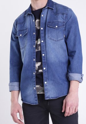 Man die een blauw denimhemd met opgestroopte mouwen draagt over een zwart-grijs camouflage t-shirt, gecombineerd met een zwarte broek.