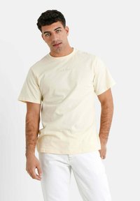 Gul bomulds T-shirt med korte ærmer, rund hals, prydet med diskret broderet tekst på brystet. Båret med hvide jeans.