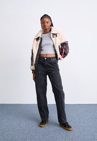 Giacca bomber in shearling marrone con maniche grigie, top corto grigio, jeans neri a vita alta con dettagli dorati e sneakers nere con suole color sabbia.