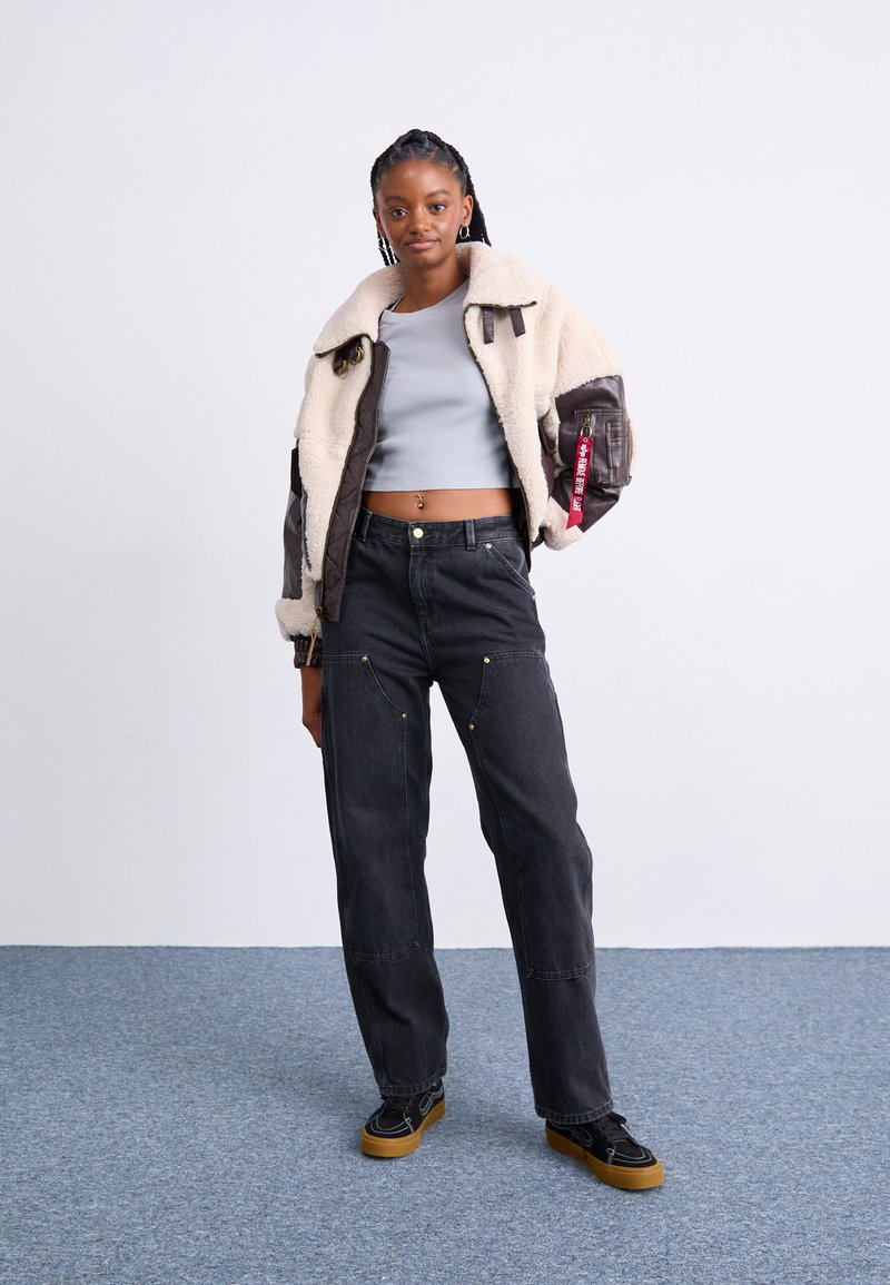 Braune Schaffell-Bomberjacke mit grauen Ärmeln, grauer Cropped-Top, hochtaillierte schwarze Jeans mit goldenen Akzenten und schwarze Sneaker mit braunen Sohlen.