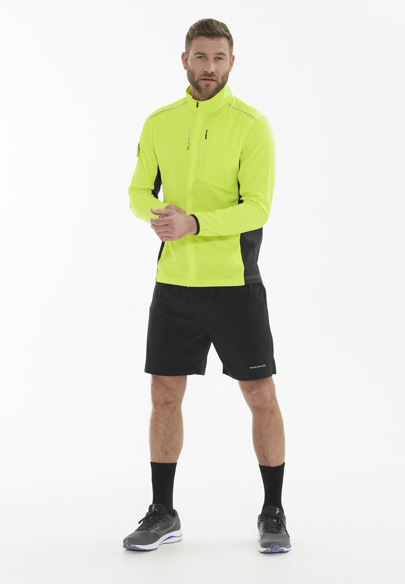 Lys gul jakke med sorte detaljer, lynlåslommer og tætsiddende design, parret med sorte shorts og grå træningssko.