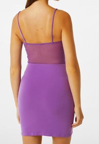 Donna che indossa un vestito viola aderente con sottili spalline e un pannello trasparente nella parte centrale della schiena, mostrata di spalle su uno sfondo bianco.