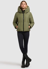 Olijfgroene pufferjack met een grote capuchon, rits aan de voorkant en elastische manchetten. Gecombineerd met zwarte leggings en stevige zwarte veterschoenen.