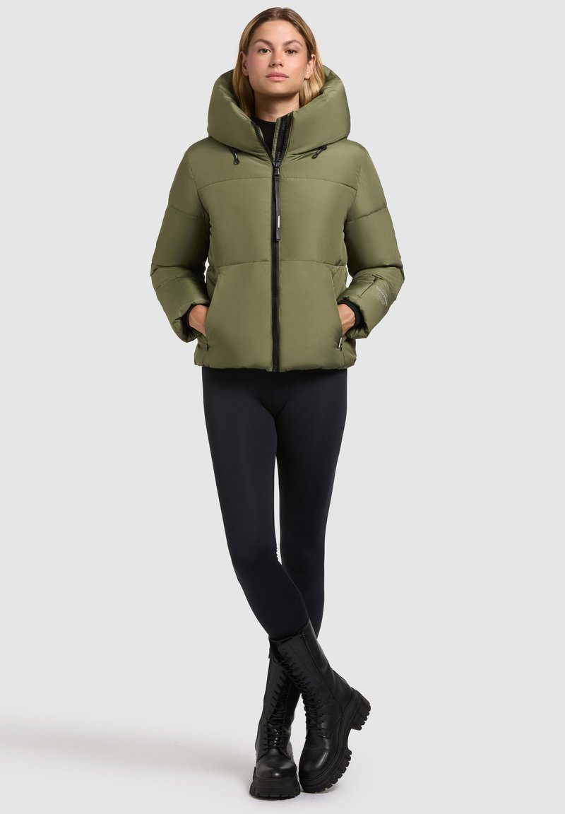 Olijfgroene pufferjack met een grote capuchon, rits aan de voorkant en elastische manchetten. Gecombineerd met zwarte leggings en stevige zwarte veterschoenen.