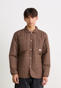 Bruine gewatteerde jas met een kraag, voorzien van twee voorzakken en drukknoopsluitingen. Glad textiel met een zichtbaar patroon van verticale lijnen.