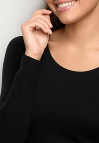 Schwarzes Langarmoberteil mit rundem Ausschnitt, glatter Stofftextur, taillierter Passform und sichtbaren Nähten. Die Hand des Models berührt das Kinn.