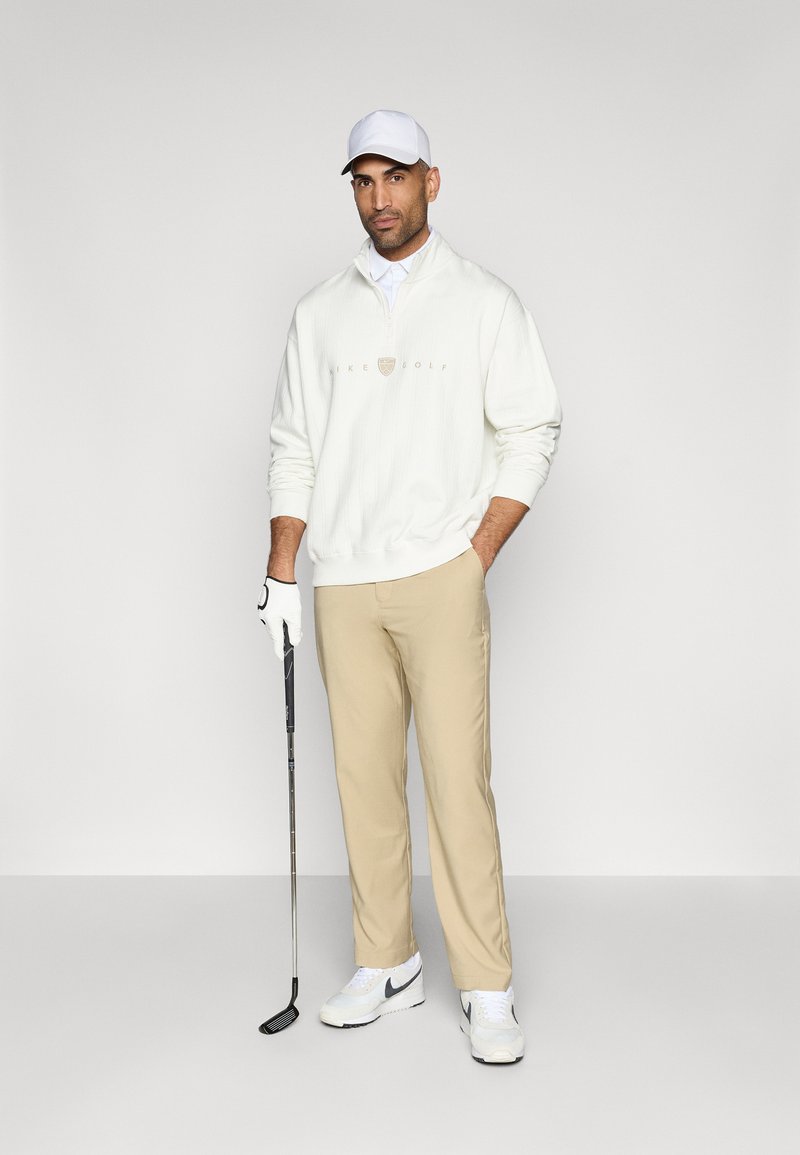 Weißer Reißverschluss-Golfsweater mit Logo, beige Golfhosen, weiße Sneakers mit grauen Akzenten und eine weiße Kappe, während er einen Golfschläger hält.