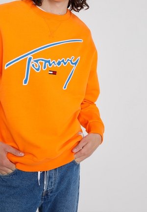 Osoba nosząca jaskrawy pomarańczowy sweter Tommy Hilfiger z niebieskim logo w stylu pisma oraz małą emblematem flagi, ubrana w niebieskie dżinsy, z rękami w kieszeniach.