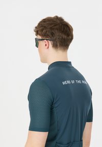 Cykeltrøje med korte ærmer i mørk teal, med tekstureret stof, en stående krave og "HERO OF THE ROAD" trykt på bagsiden.