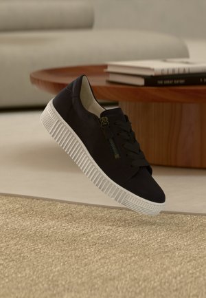 Schwarzer Sneaker mit weißer dicker Sohle und seitlichem Reißverschluss schwebt über beigem Teppich, runder Holztisch mit gestapelten Büchern im Hintergrund.