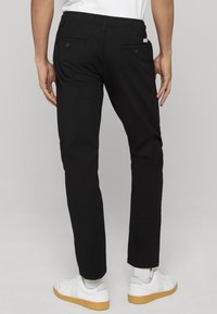 Pantalon noir à coupe ajustée, doté de poches arrière. Porté avec des baskets blanches, affichant un style décontracté. Tissu doux et design simple.