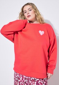 Sweatshirt rouge surdimensionné avec des épaules tombantes, présentant un graphique en forme de cœur avec le texte "DON'T" en rose. Tissu doux et texturé.
