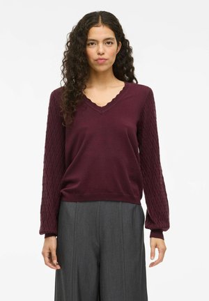 Pull en tricot bordeaux avec un col en V festonné, des manches texturées et une coupe décontractée, associé à un pantalon gris ajusté.