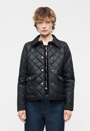 Rövid hajú nő fekete steppelt kabátot visel patent gombokkal, fekete felső és kék farmer nadrág alatt, egyszerű háttér előtt áll.