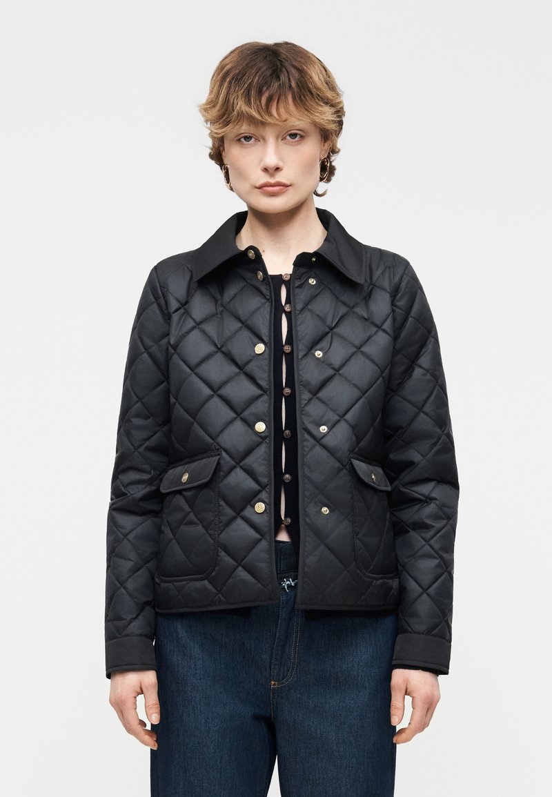 Γυναίκα με κοντά μαλλιά που φοράει μαύρο μπουφάν με καπιτονέ και κουμπιά τύπου σναπ πάνω από μαύρο τοπ και μπλε τζιν, που στέκεται μπροστά σε απλό φόντο.