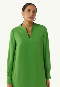Grünes, langärmeliges Kleid mit V-Ausschnitt und lockerer Passform. Hergestellt aus glattem Stoff mit leichtem Glanz; verfügt über geraffte Bündchen und minimale Details.