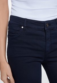 Primer plano de una persona que lleva vaqueros azul oscuro con cierre de botón y cremallera, y una blusa blanca metida por dentro.