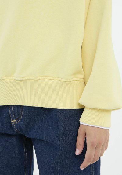 Gul bomuldsstriktrøje med ribbestrikkede manchetter og nederste kant. Har en afslappet pasform og en subtil tekstur, parret med mørkeblå denimjeans.