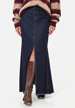 Donna che indossa una lunga gonna di jeans scuro con spacco frontale, stivali marroni al ginocchio con punte dorate e un maglione a righe soffice.