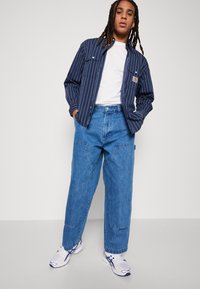 Navy striped jacket with two chest pockets, white t-shirt, light blue baggy denim jeans, and white sneakers with blue accents.