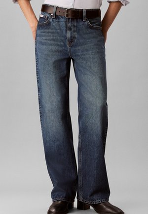 Person iført mørkeblå jeans med lige ben, brun læderbælte, hvid skjorte stukket ind og brune lædersko. Hænderne i lommerne.