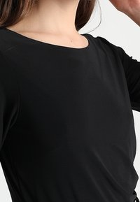 Černý top s kulatým výstřihem a tříčtvrtečními rukávy, vyrobený z hladké tkaniny s jemným záhybem.