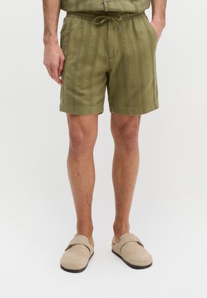 Man draagt olijfgroene textuurshorts met koordsluiting en beige instappers, staand tegen een effen achtergrond.