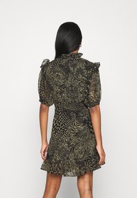 Kvinna med långt mörkt hår iklädd en kort, volangklädd klänning med puffärmar och blandade djur- och blommönster, som står mot en enkel bakgrund.