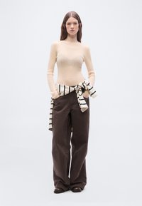 Femme portant un haut beige à manches longues côtelées, pantalon large marron foncé, chaussures noires, avec un pull rayé noué autour de la taille, mains dans les poches.